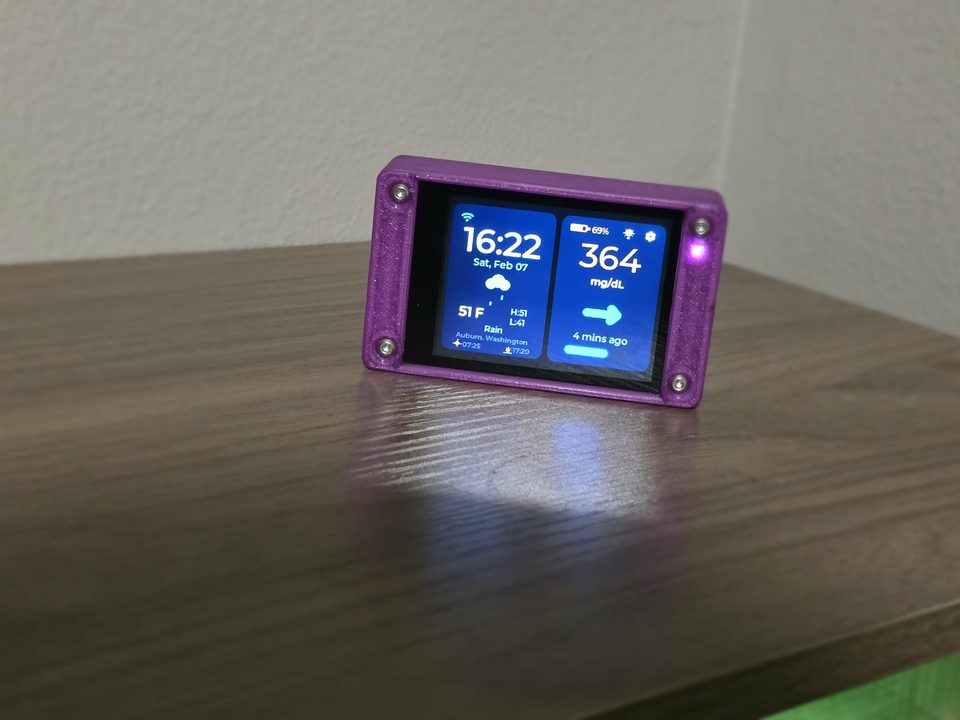 An assembled CYGM unit on a desk in a printed case, showing a glucose reading, a trend arrow and the time.