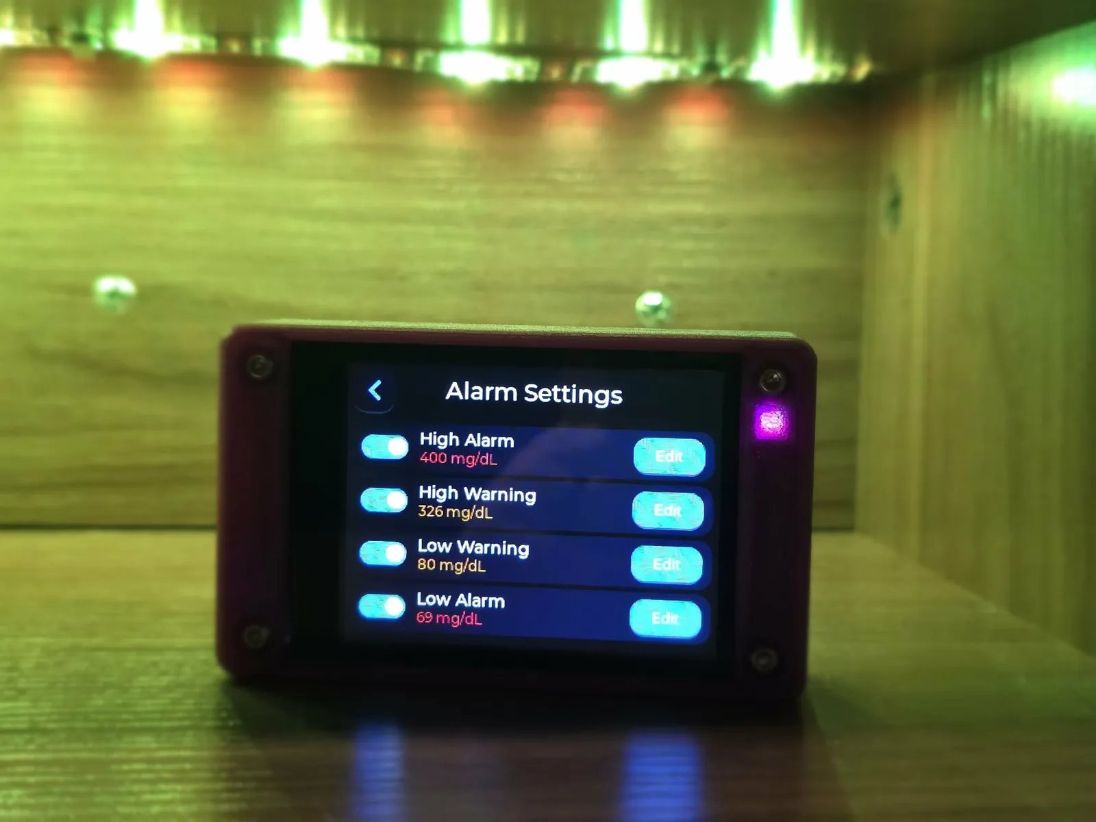 A purple printed case sitting on a wooden shelf under warm green lighting, its screen showing the Alarm Settings list with High Alarm, High Warning, Low Warning and Low Alarm rows, each with a toggle and an Edit button.