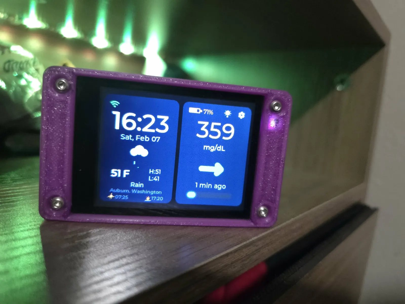A purple printed case angled on a dark wooden shelf edge under green accent lighting, its home screen showing 359 mg/dL with a flat side arrow beside the time, weather and location.