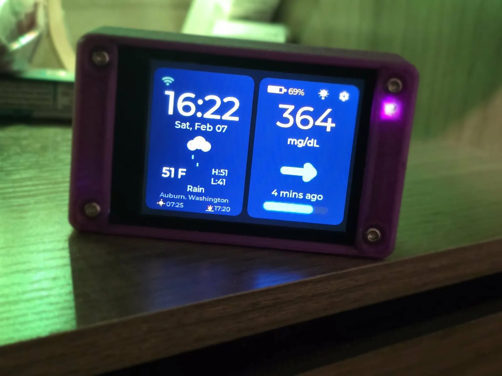 A purple printed case in close three-quarter view on a wooden surface washed in green light. The home screen reads 16:22 Sat, Feb 07 with a rain icon, 51 F and Auburn, Washington on the left, and 364 mg/dL with a flat side arrow, 4 mins ago and a 69% battery on the right.