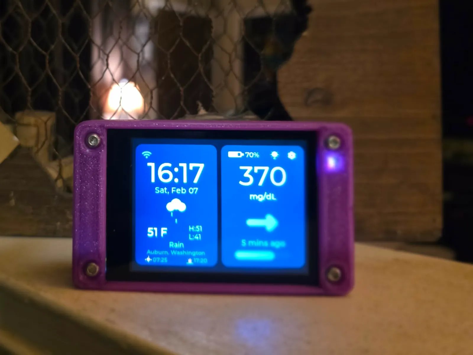 A purple printed case standing on a light wooden ledge in front of a fireplace, its split home screen showing 370 mg/dL with a flat side arrow on the right and the time, weather and location on the left.