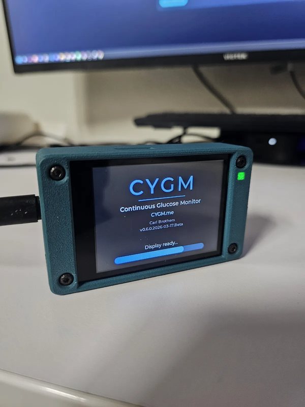 A CYGM in a teal printed case standing on a white desk in front of a monitor, showing the boot splash with the CYGM wordmark and a Display ready progress bar.