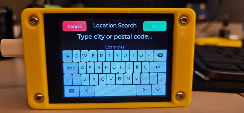 A CYGM in a yellow printed case on a desk with a USB cable attached, showing the location search screen with a Type city or postal code field above an on-screen keyboard.
